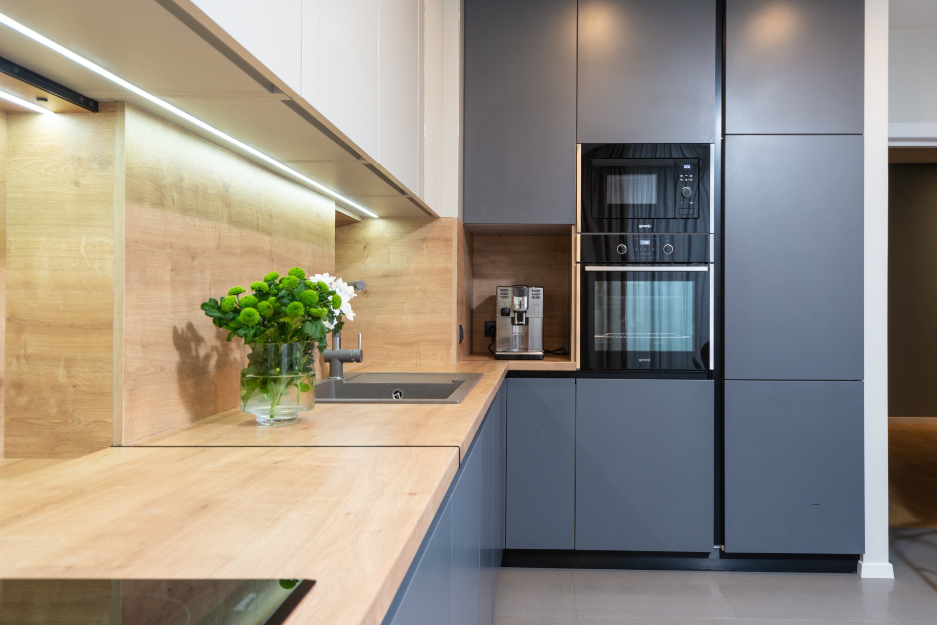 Nowoczesna kuchnia z drewnianym blatem, szarymi szafkami i wbudowanym sprzętem AGD. Na blacie wazon z kwiatami i ekspres do kawy.