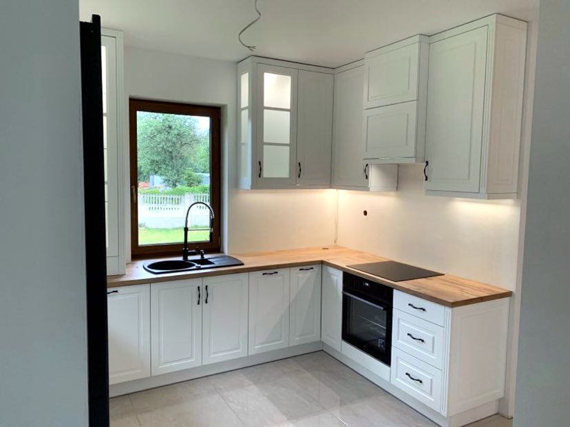 Jasna kuchnia z drewnianym blatem, czarnym zlewem i oknem z widokiem na zieleń. Górne szafki częściowo przeszklone, oświetlenie LED pod szafkami.