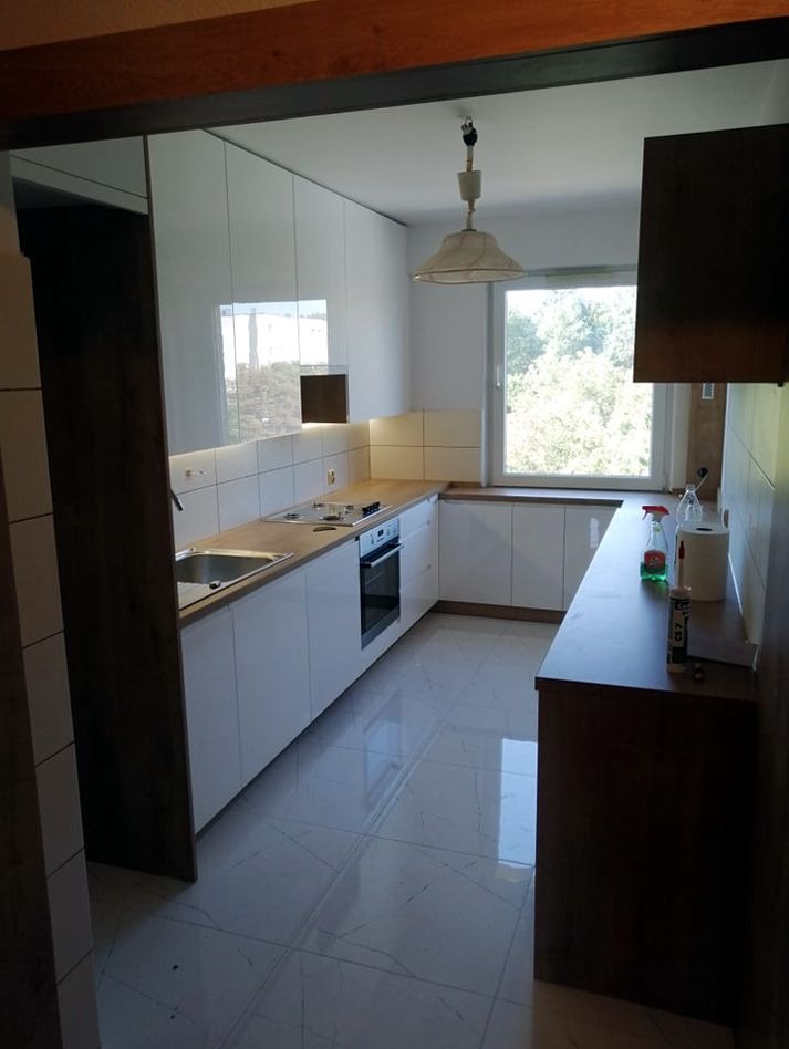 Biała kuchnia z połyskiem, drewniany blat, płytki na ścianie i podłodze. Widok na okno i zieleń za nim. Całość w nowoczesnym stylu.