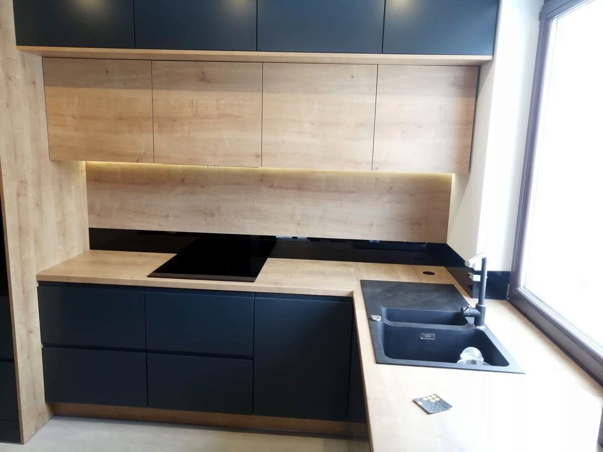 Nowoczesna kuchnia z drewnianymi szafkami górnymi i granatowymi dolnymi, z czarnym blatem i płytą indukcyjną. Oświetlenie LED pod szafkami dodaje ciepła.