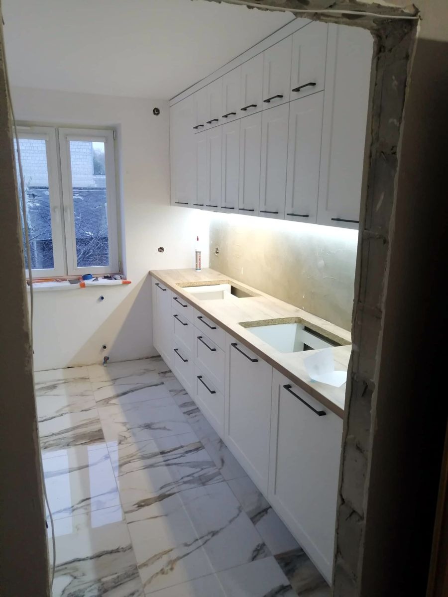 Biała kuchnia z czarnymi uchwytami, marmurowe płytki podłogowe, widok przez surowy otwór drzwiowy. Częściowo zamontowana, oświetlenie podszafkowe.