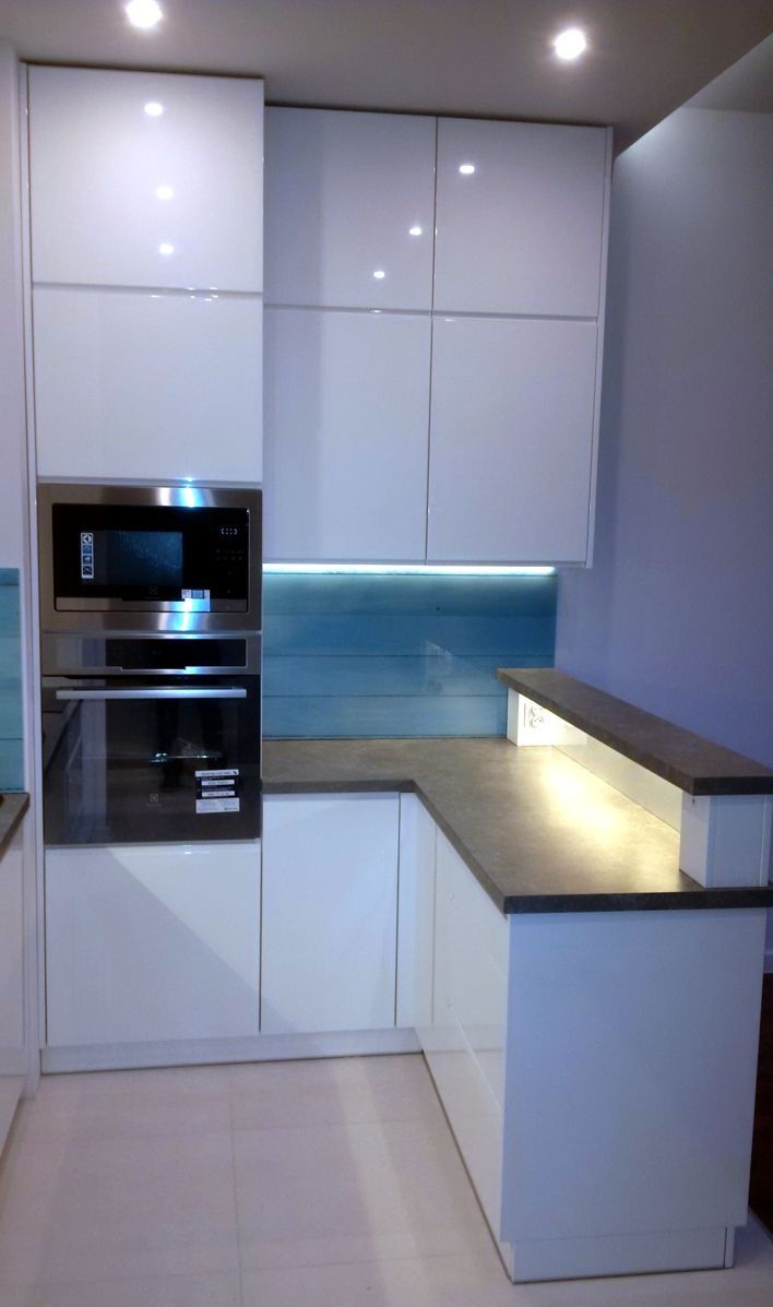 Biała, nowoczesna kuchnia z zabudową pod sufit, z piekarnikiem i mikrofalówką w zabudowie. Blat w kolorze szarości, podświetlony panel nad blatem.