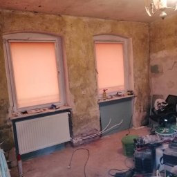 Majstersztyk - Pokój w trakcie remontu: ściany z resztkami starej farby, dwa okna z roletami, grzejnik, narzędzia i materiały budowlane na podłodze. Widoczna instalacja grzewcza przy ścianie.