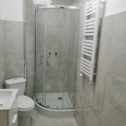 Majstersztyk - Wykończona łazienka w szarościach z kabiną prysznicową z zaokrąglonym frontem, grzejnikiem drabinkowym i umywalką z szafką. Ściany i podłoga wyłożone płytkami.