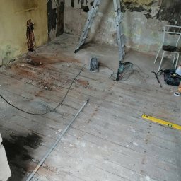 Majstersztyk - Pokój w trakcie remontu: zniszczone ściany, drewniana podłoga, drabina, narzędzia (wiertarka, poziomica). Widoczne ślady po instalacjach i przygotowania do prac wykończeniowych.
