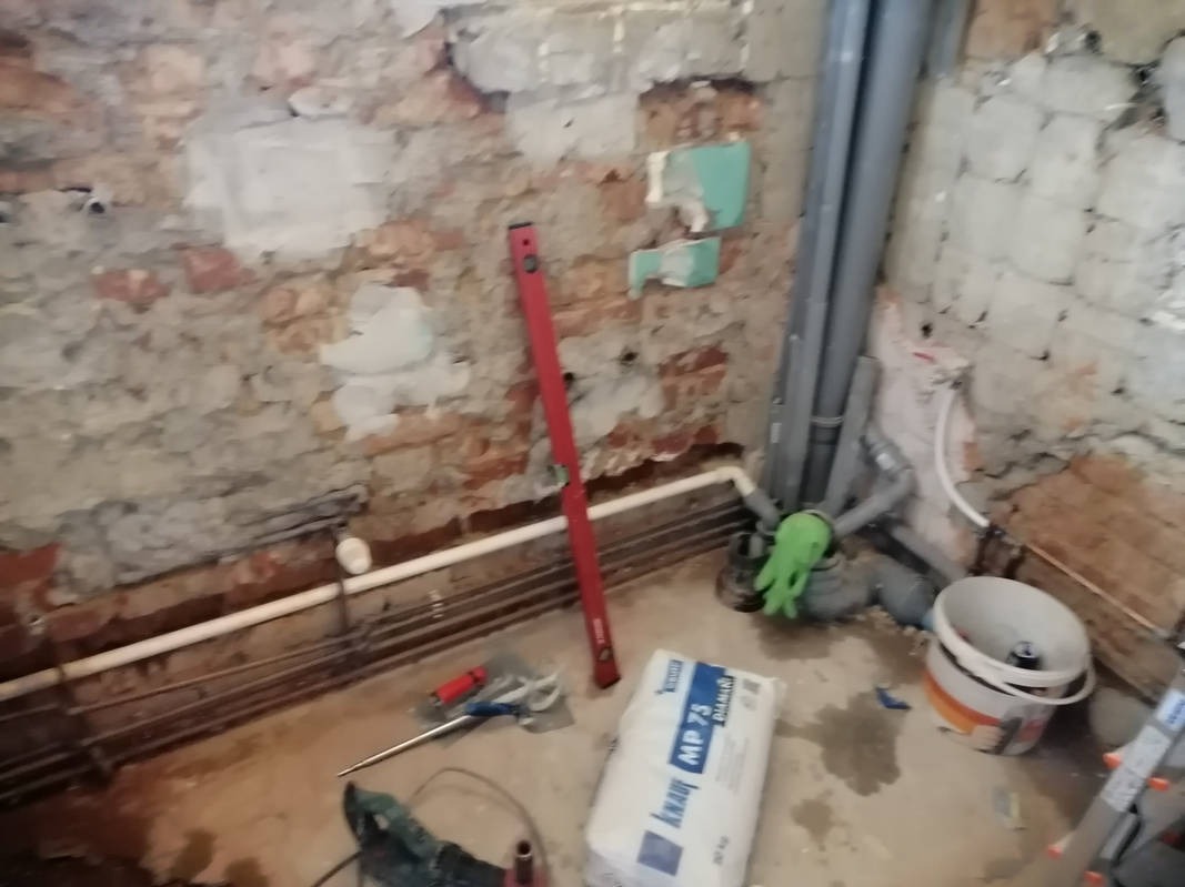 Wnętrze w trakcie remontu: odsłonięta cegła, rury, poziomica, worek gładzi Knauf MP 75, narzędzia i wiadro. Widoczne ślady prac remontowych.