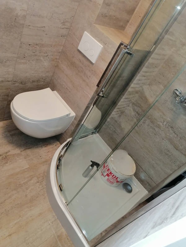 Nowoczesna łazienka po remoncie: podwieszana toaleta, kabina prysznicowa z widocznym wiadrem po kleju, beżowe płytki imitujące kamień na ścianach i podłodze.