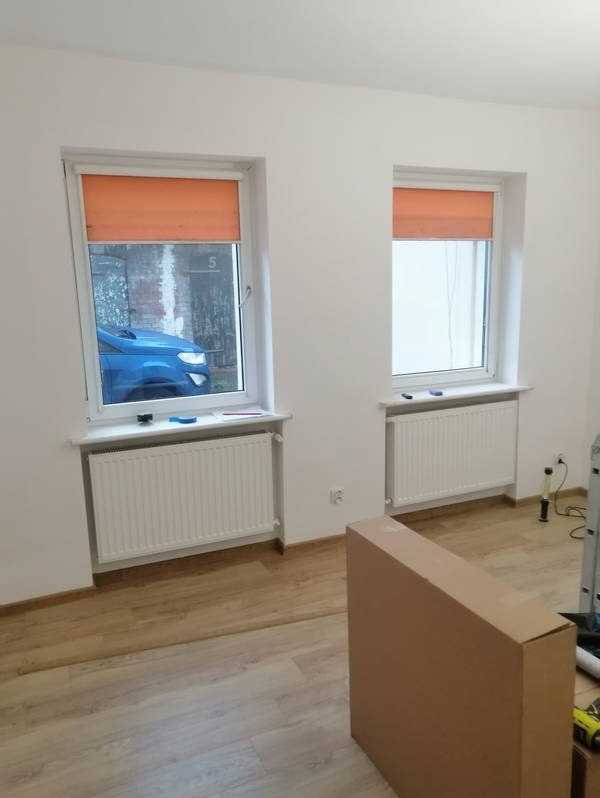 Wnętrze pokoju po malowaniu ścian na biało, z dwoma oknami z pomarańczowymi roletami i grzejnikami. Na podłodze panele, w tle kartonowe pudło.