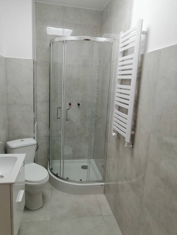 Wykończona łazienka w szarościach z kabiną prysznicową z zaokrąglonym frontem, grzejnikiem drabinkowym i umywalką z szafką. Ściany i podłoga wyłożone płytkami.