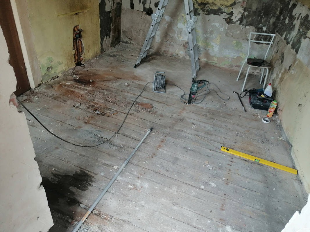 Pokój w trakcie remontu: zniszczone ściany, drewniana podłoga, drabina, narzędzia (wiertarka, poziomica). Widoczne ślady po instalacjach i przygotowania do prac wykończeniowych.