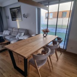 Drewniane Klimaty - Stół z blatem z jasnego drewna i czarnymi, metalowymi nogami w kształcie litery V. Na stole wazon z kwiatami, w tle okno z żaluzjami i część salonu.