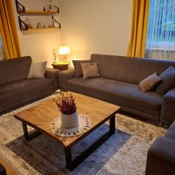 Drewniane Klimaty - Salon z brązowymi sofami i drewnianym stolikiem kawowym z kwiatami. Żółte zasłony w oknach i półki z dekoracjami na ścianie tworzą przytulną atmosferę.