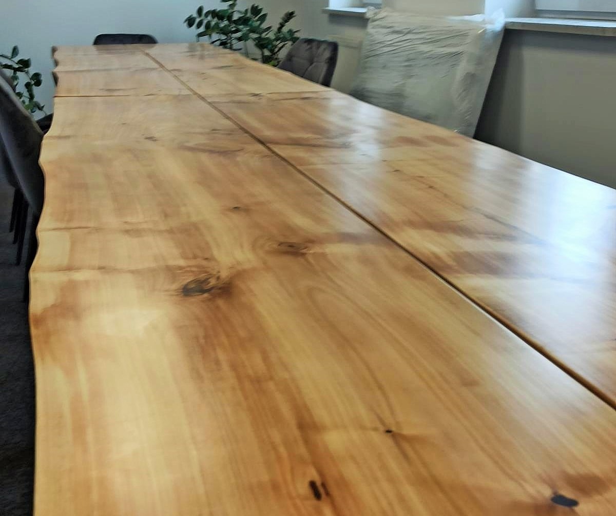 Długi, jasny blat drewniany z naturalnymi krawędziami, złożony z kilku części. Widoczne sęki i unikalny wzór drewna. W tle krzesła i roślina doniczkowa.