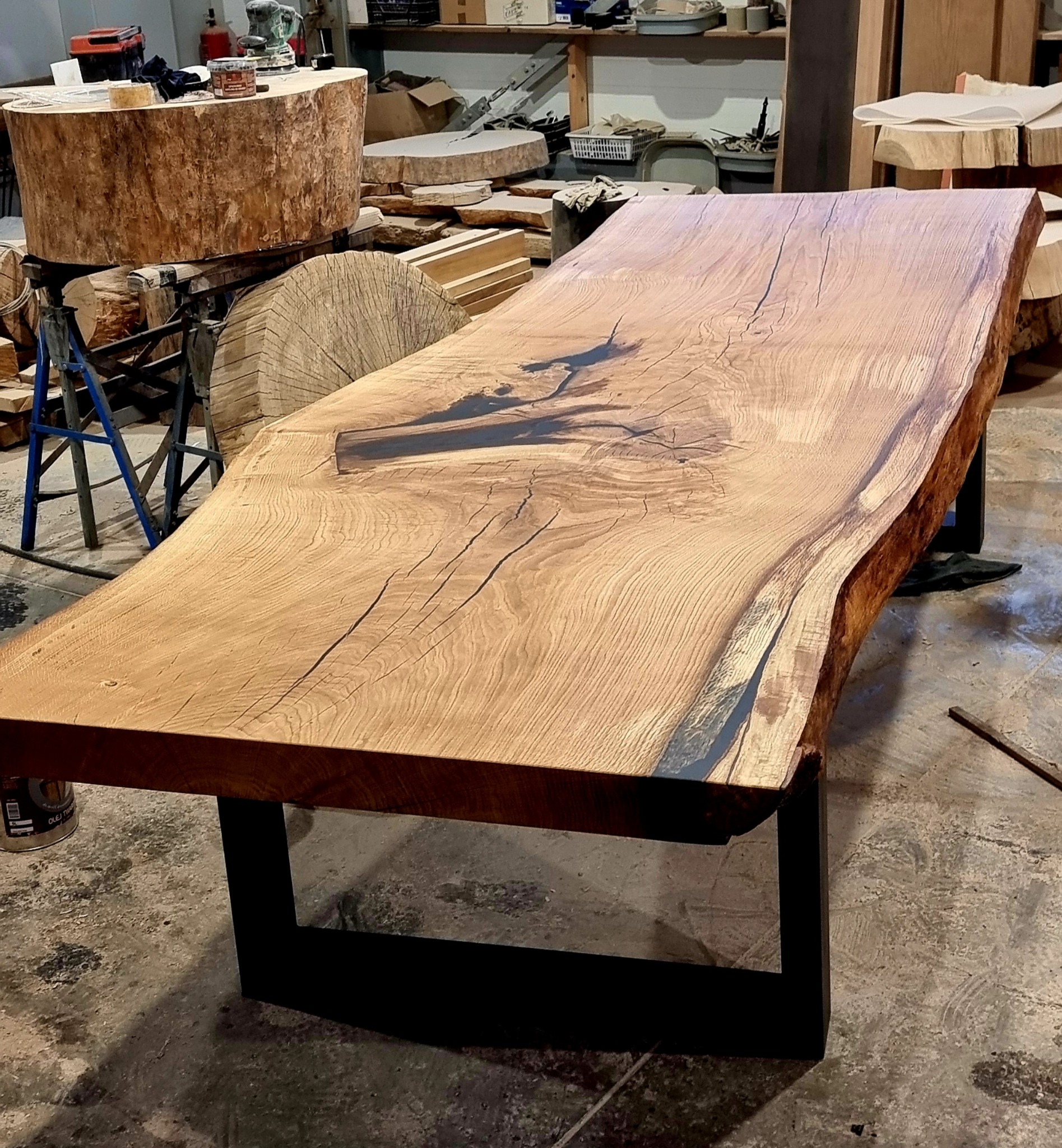 Wyjątkowy blat drewniany z żywicą epoksydową, osadzony na metalowych nogach. Widoczne słoje drewna i naturalne krawędzie. W tle warsztat stolarski.