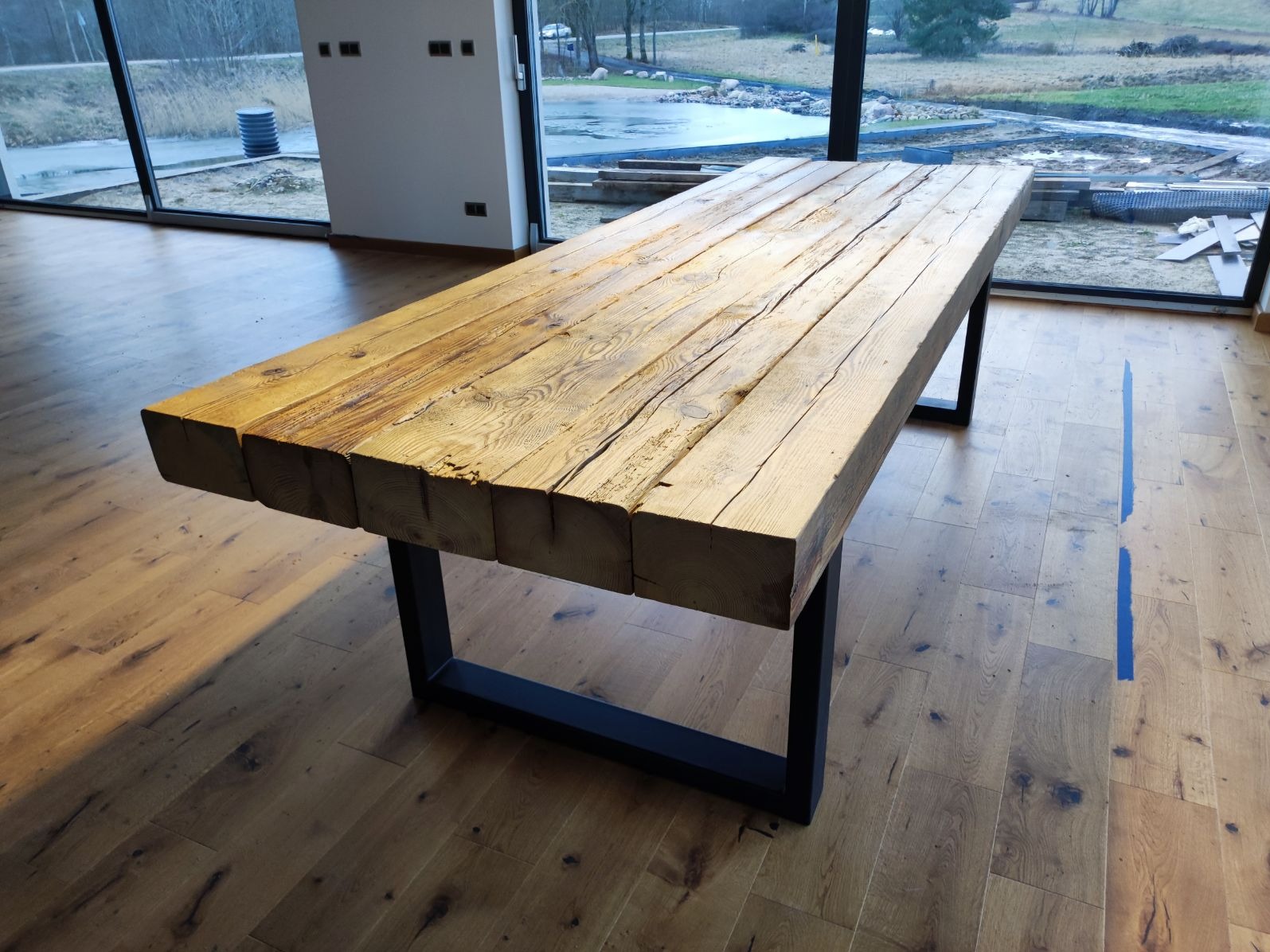 Rustykalny stół z grubych, drewnianych belek na metalowych nogach w nowoczesnym wnętrzu z dużymi oknami i drewnianą podłogą.