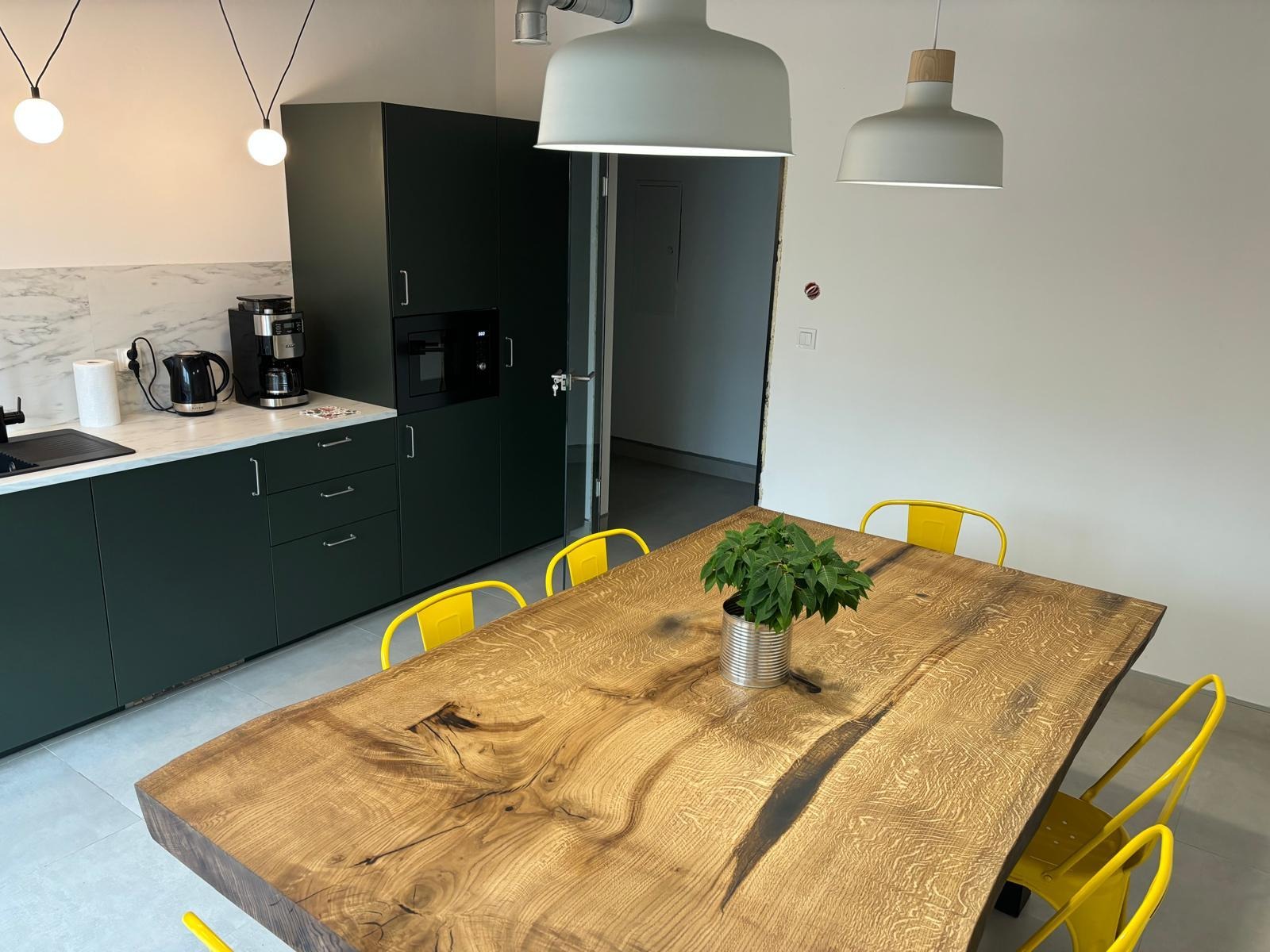 Wnętrze kuchni z ciemnozielonymi szafkami i blatem z marmuru. Na pierwszym planie duży, drewniany stół z żółtymi krzesłami i rośliną w puszce. Nowoczesne lampy wiszące.