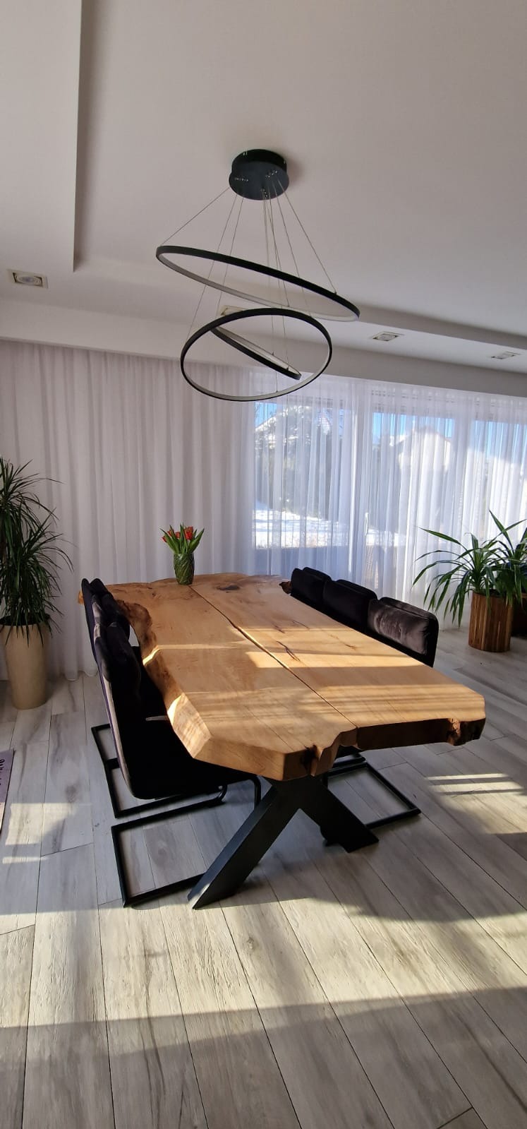 Stół z blatem z litego drewna o nieregularnych krawędziach, na metalowej podstawie w kształcie litery X. Nad stołem nowoczesna lampa z okręgów.