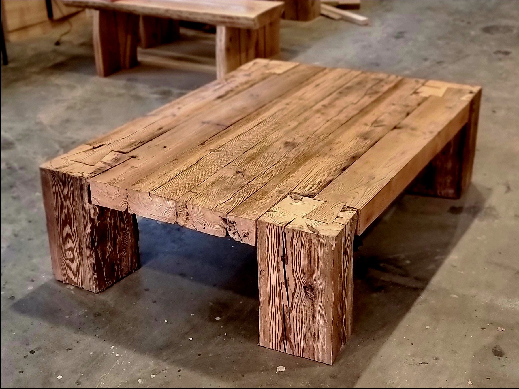 Rustykalny stolik kawowy z grubych desek i nóg z litego drewna, widoczny z bliska, z innym stolikiem w tle, ukazujący unikalną strukturę drewna.