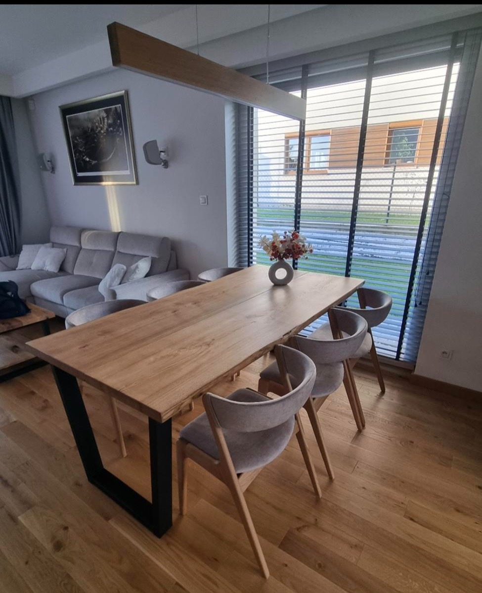 Stół z blatem z jasnego drewna i czarnymi, metalowymi nogami w kształcie litery V. Na stole wazon z kwiatami, w tle okno z żaluzjami i część salonu.