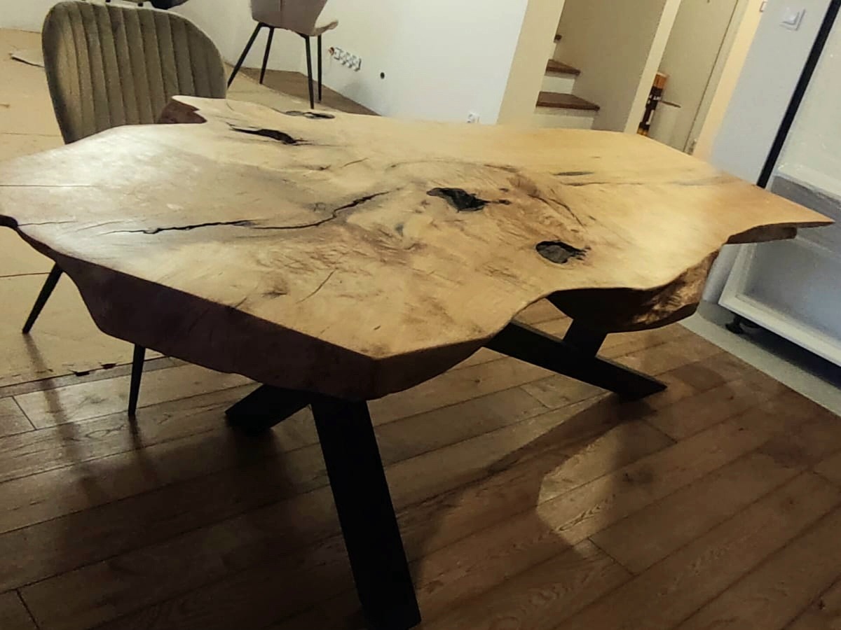 Unikatowy stół z blatem z litego drewna o nieregularnym kształcie, wsparty na czarnych, metalowych nogach. Widoczne słoje i naturalne pęknięcia drewna.