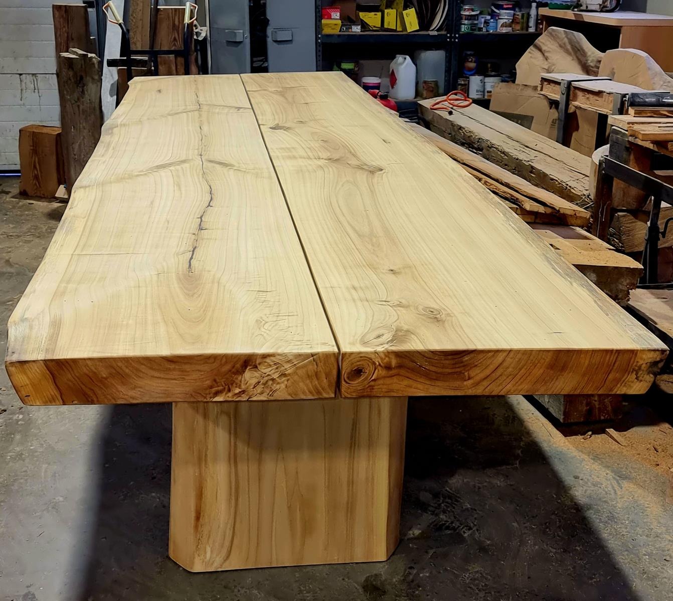 Stół z dwóch blatów z jasnego drewna o nieregularnych krawędziach, wsparty na solidnej, drewnianej podstawie. Widoczne słoje i naturalne pęknięcia drewna.