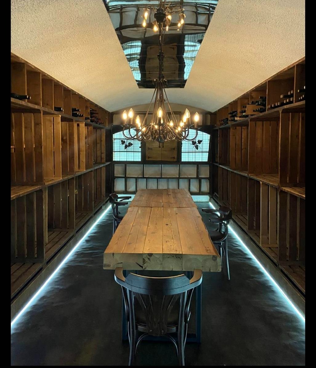 Wnętrze winiarni z drewnianymi regałami na wino, długim stołem z surowego drewna i dekoracyjnym żyrandolem pod lustrzanym sufitem. Podświetlenie podłogowe LED.