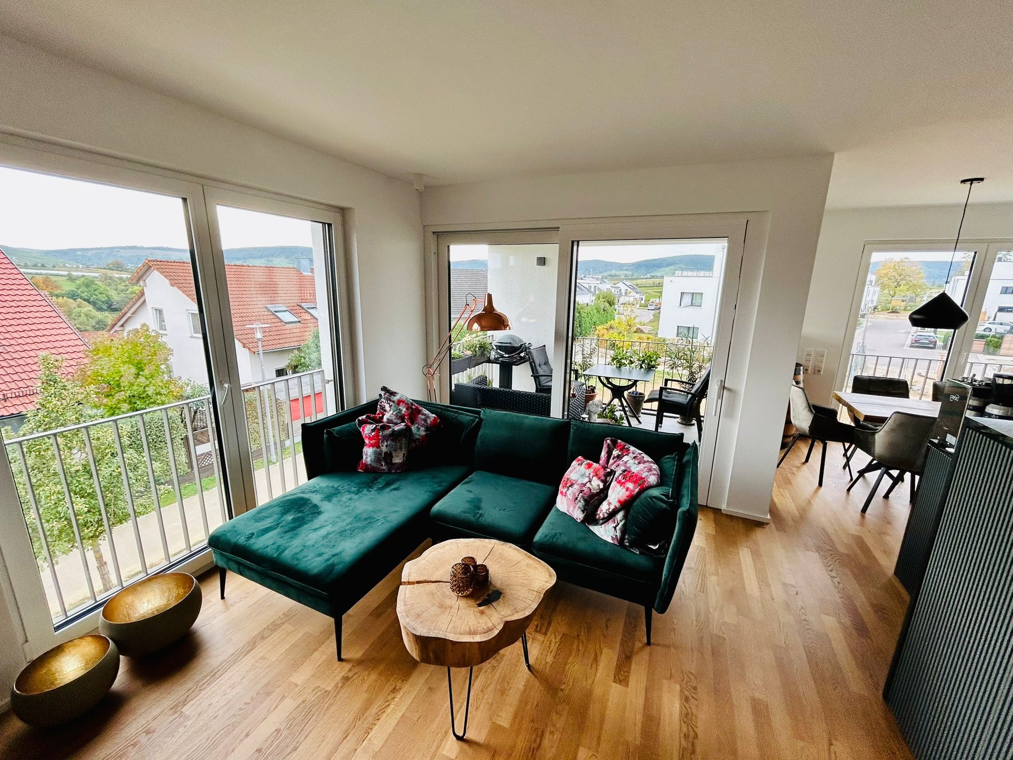 Nowoczesny salon z zieloną sofą narożną, drewnianym stolikiem kawowym i widokiem na balkon z zabudową. Jasne wnętrze z parkietem i złotymi misami dekoracyjnymi.