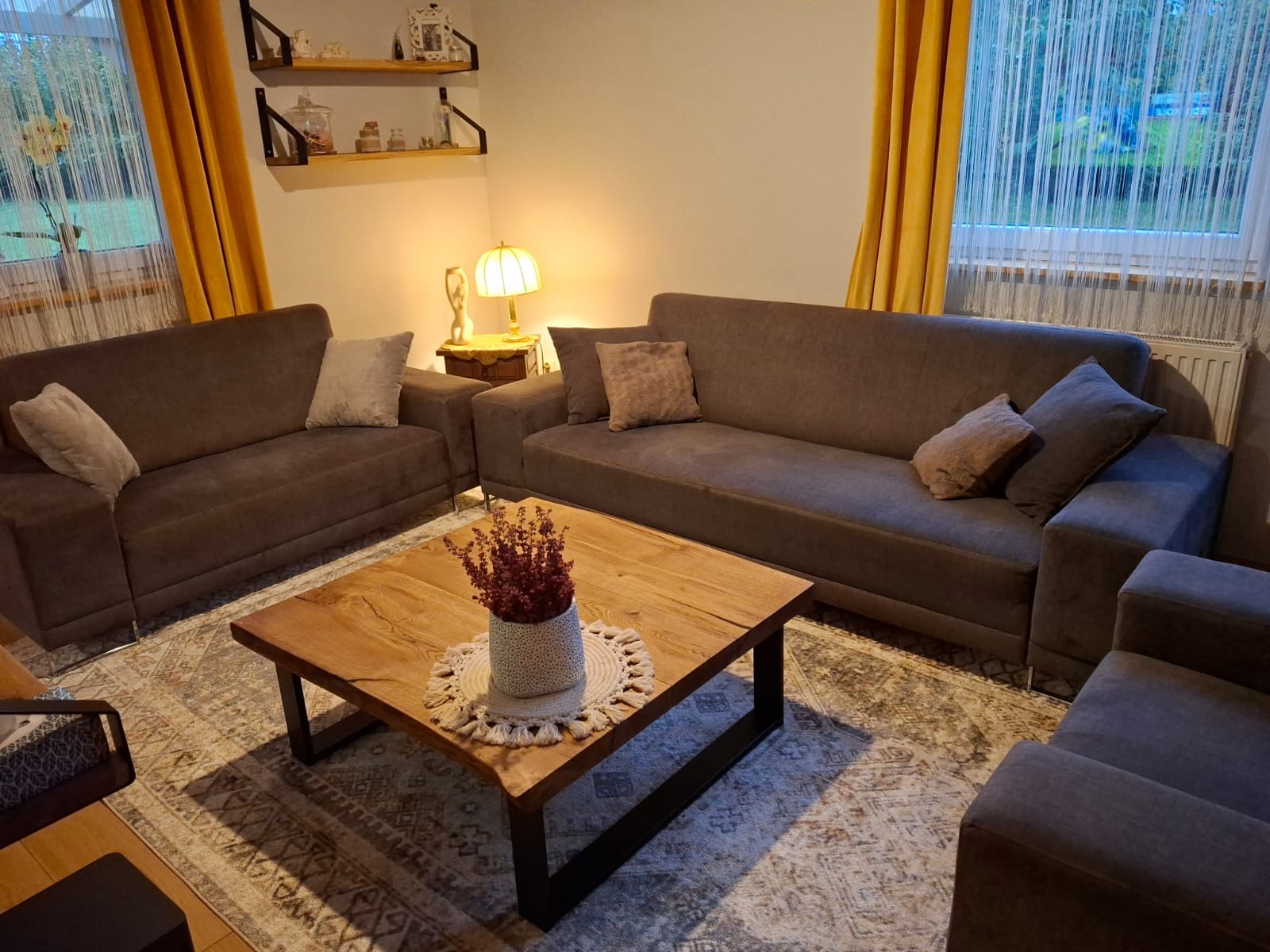 Salon z brązowymi sofami i drewnianym stolikiem kawowym z kwiatami. Żółte zasłony w oknach i półki z dekoracjami na ścianie tworzą przytulną atmosferę.