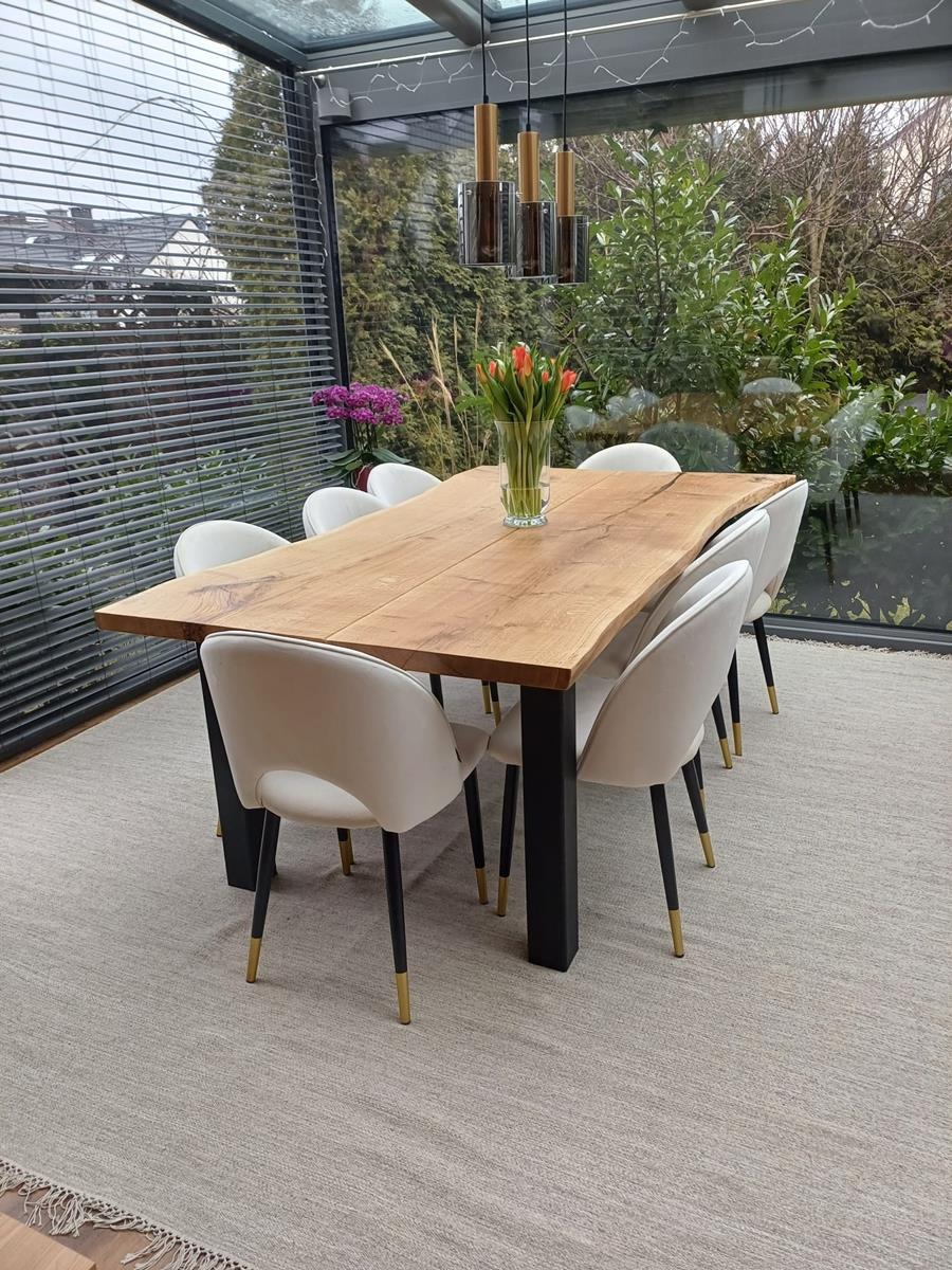 Stół z blatem z litego drewna o nieregularnych krawędziach, na czarnych nogach, otoczony białymi krzesłami. Nad nim wiszą lampy w szklanych kloszach. Tło: przeszklona weranda.