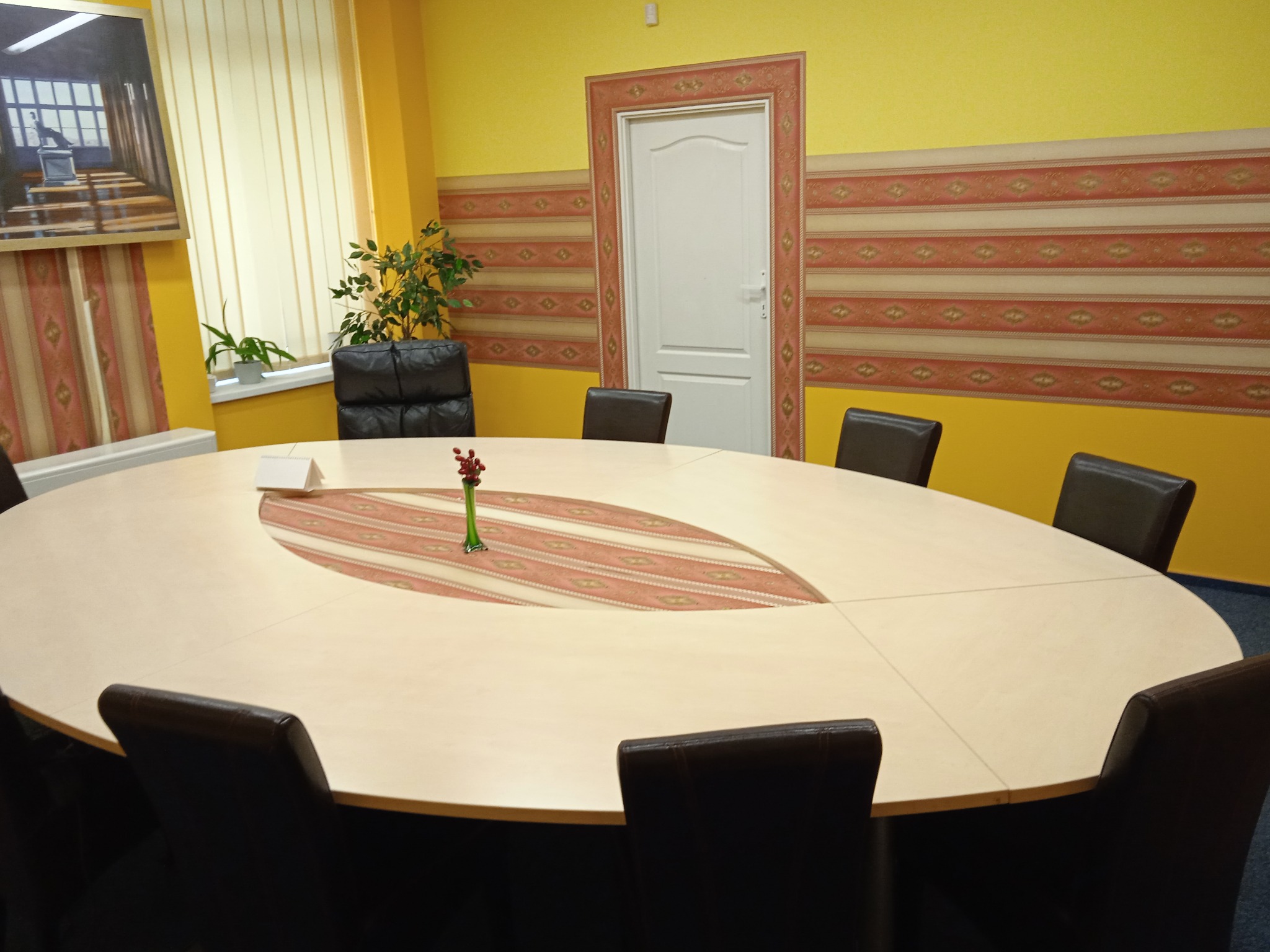 Jasna sala konferencyjna z owalnym stołem i krzesłami. Na ścianach żółta farba i dekoracyjny pas z wzorem. Na stole wazon z kwiatami.