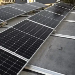 Rząd paneli fotowoltaicznych, zamontowanych na płaskim dachu, widocznych pod kątem. Panele ułożone na metalowej konstrukcji, z widocznymi elementami mocującymi.