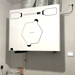 Budotom Pompy Ciepła - Nowoczesny rekuperator Thessla Green zamontowany w pomieszczeniu technicznym, z widocznymi przewodami wentylacyjnymi i panelem sterowania na ścianie.