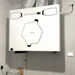 Nowoczesny rekuperator Thessla Green zamontowany w pomieszczeniu technicznym, z widocznymi przewodami wentylacyjnymi i panelem sterowania na ścianie.