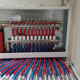 Budotom Pompy Ciepła - Rozdzielacz ogrzewania podłogowego z rurami w kolorach czerwonym i niebieskim, zamontowany w ścianie, na siatce grzewczej. Widoczne zawory i etykiety.