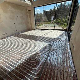 Budotom Pompy Ciepła - Białe rury ogrzewania podłogowego ułożone na folii aluminiowej w surowym wnętrzu z dużym oknem i widokiem na zieleń. Równoległe ułożenie rur.