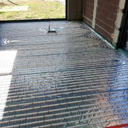 Budotom Pompy Ciepła - Ułożone rury ogrzewania podłogowego na folii izolacyjnej w surowym wnętrzu, widoczne elementy instalacji elektrycznej na ścianie z cegły.