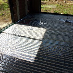 Budotom Pompy Ciepła - Białe rury ogrzewania podłogowego ułożone na srebrnej folii izolacyjnej w surowym wnętrzu budynku z widokiem na zewnątrz przez okno.