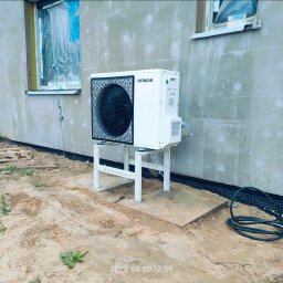 Budotom Pompy Ciepła - Biała pompa ciepła Hitachi na metalowym stelażu, zainstalowana przy ścianie budynku w trakcie budowy, z widocznym oknem zabezpieczonym folią.