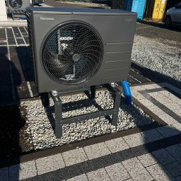 Budotom Pompy Ciepła - Szara pompa ciepła Panasonic Aquarea na metalowej konstrukcji, umieszczona na żwirowym podłożu z kostką brukową. Widoczne niebieskie rury odprowadzające.