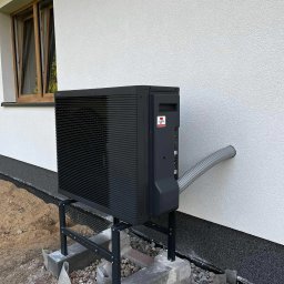 Budotom Pompy Ciepła - Czarna pompa ciepła zamontowana na zewnątrz budynku z widocznym przewodem wchodzącym w ścianę. Widoczne okno na górze i fragment fundamentu.