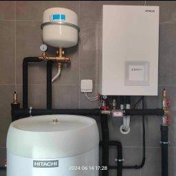 Budotom Pompy Ciepła - Zestaw urządzeń Hitachi: pompa ciepła z zbiornikiem buforowym i naczyniem wzbiorczym, instalacja w szarym pomieszczeniu, widoczne rury i zawory.