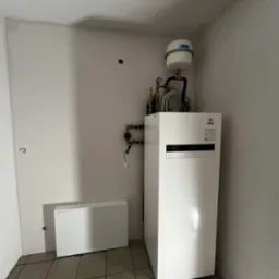 Biała pompa ciepła Panasonic Aquarea w rogu pomieszczenia z płytkami. Widoczne rury, manometry i zbiornik wyrównawczy. Obok biała szafka instalacyjna.