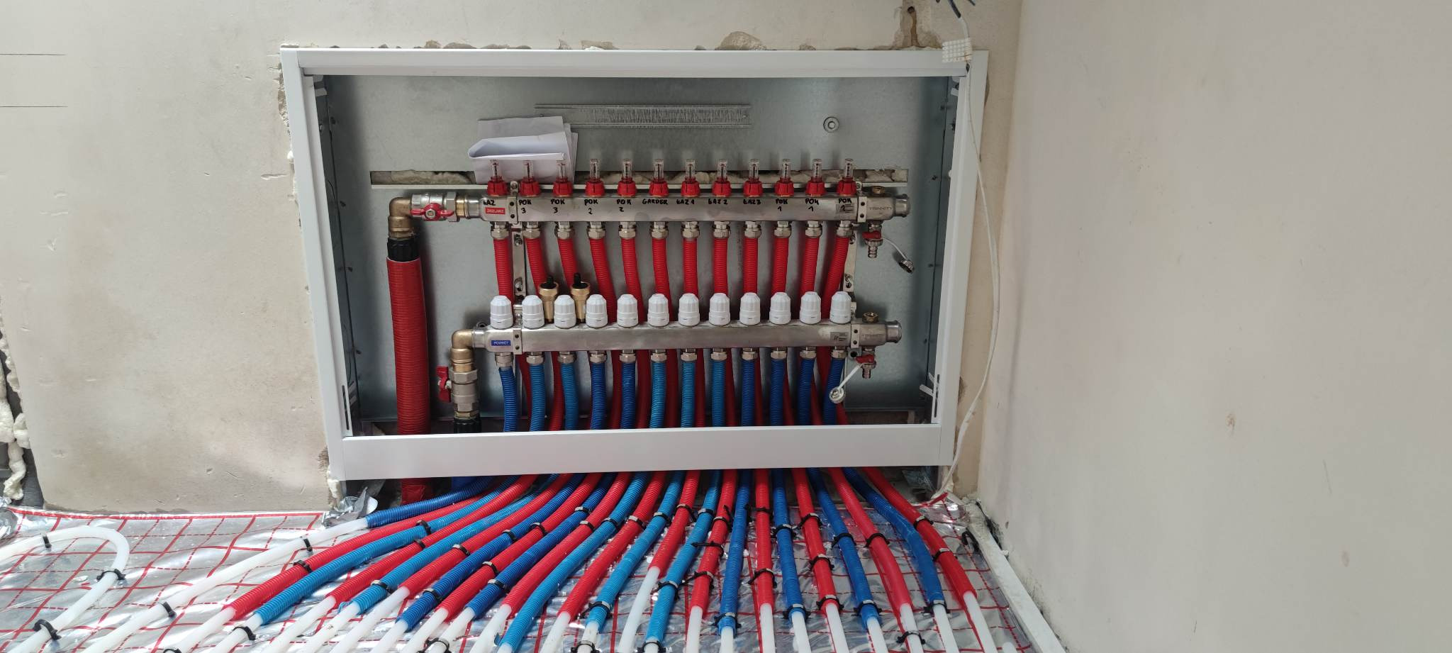 Rozdzielacz ogrzewania podłogowego z rurami w kolorach czerwonym i niebieskim, zamontowany w ścianie, na siatce grzewczej. Widoczne zawory i etykiety.