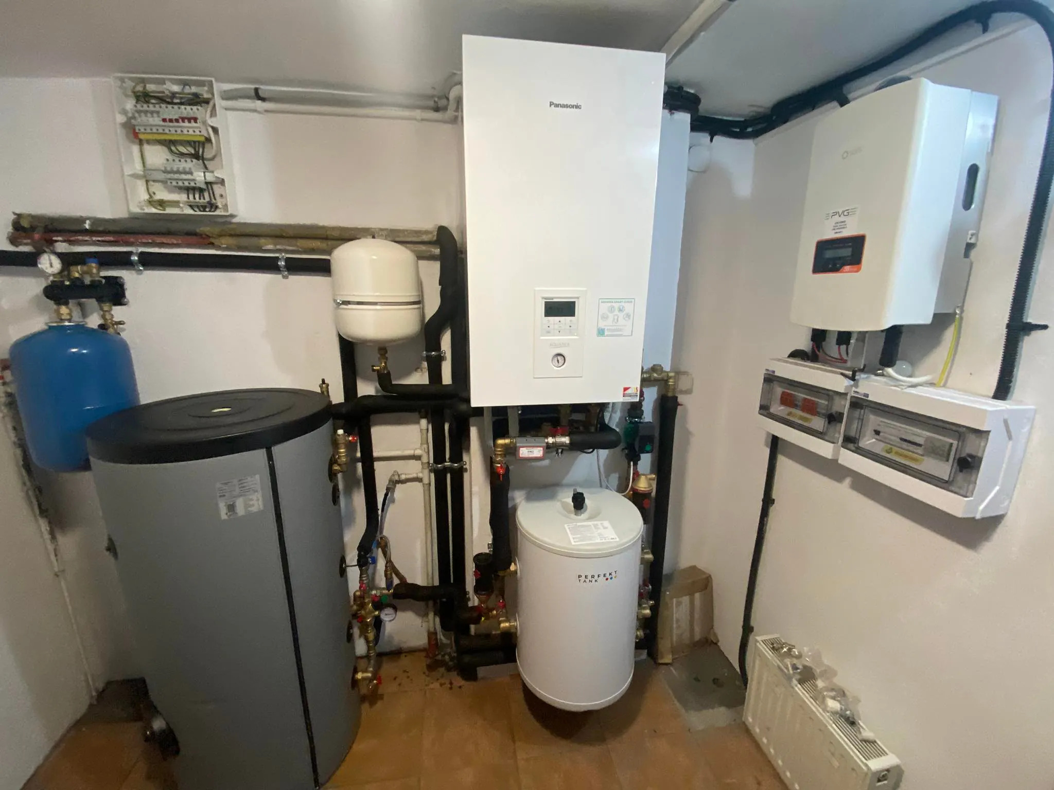 Kompletna instalacja grzewcza z pompą ciepła Panasonic, zbiornikami i inwerterem PVGE w kotłowni. Widoczne rury, zawory i rozdzielnica elektryczna.