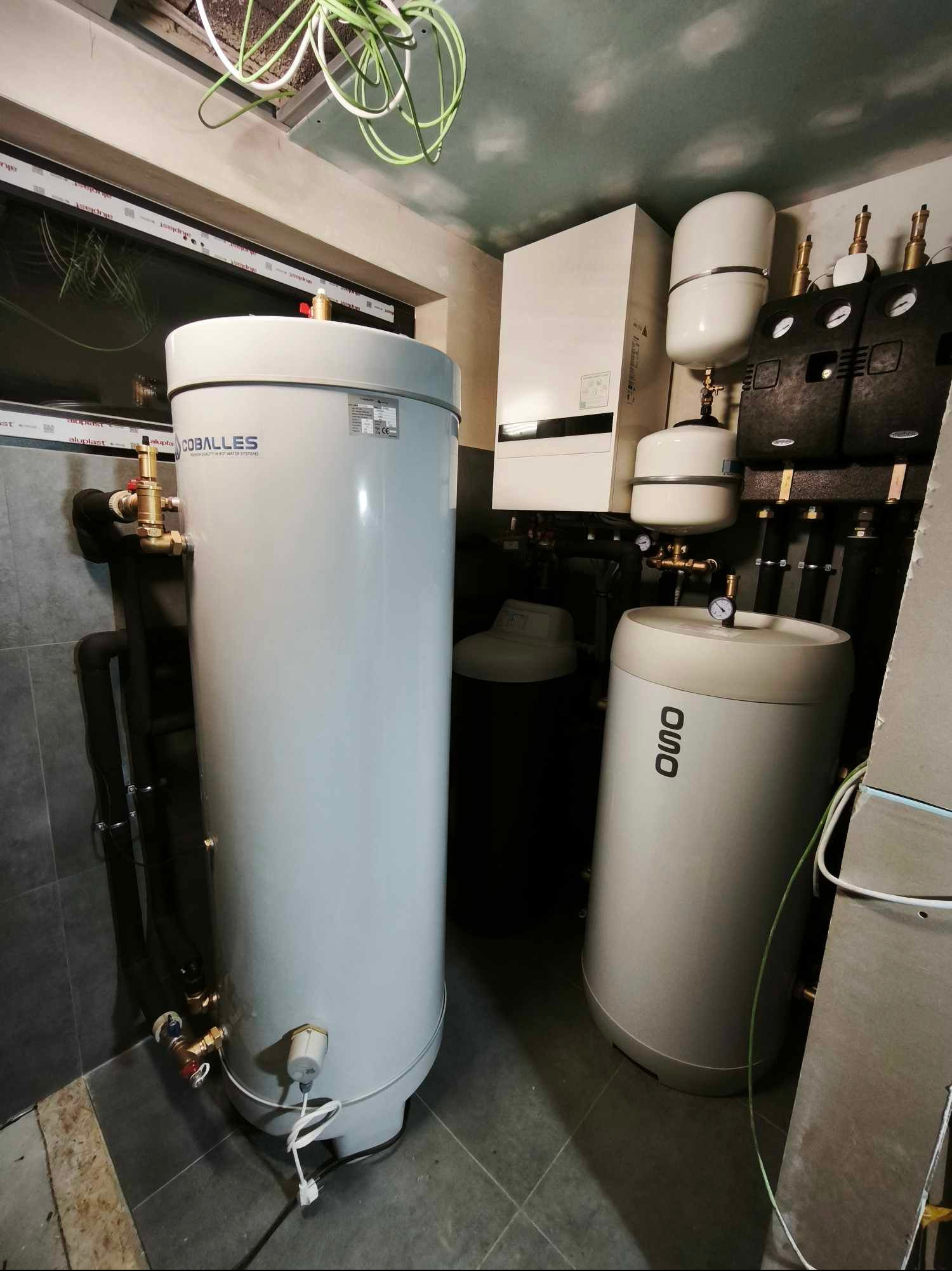 Kompletna kotłownia z dwoma zbiornikami, piecem i osprzętem na ścianie. Widoczne rury, zawory i przewody. Szare płytki na podłodze i okno w tle.