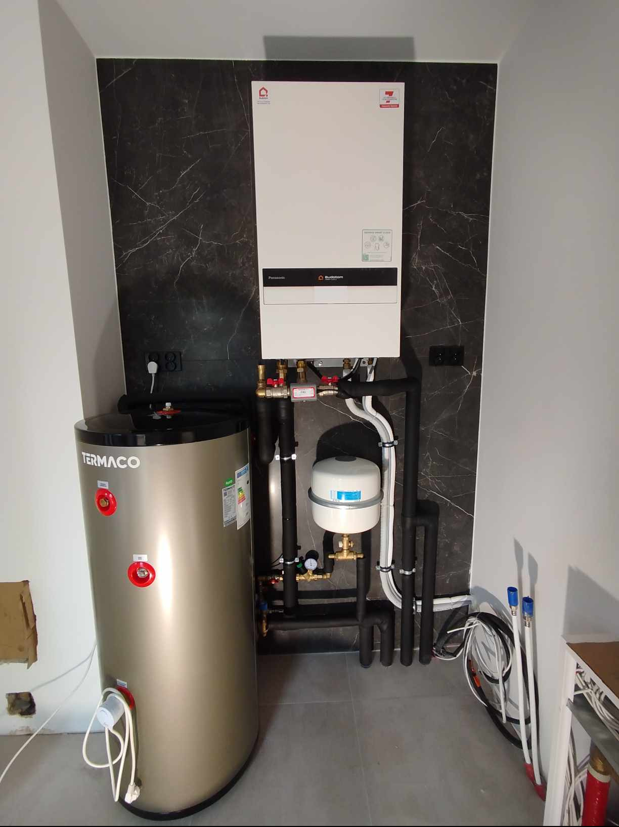 Nowoczesna instalacja grzewcza z zasobnikiem Termaco i kotłem Buderus, umieszczona na tle ściany z czarnym marmurem, widok z przodu.