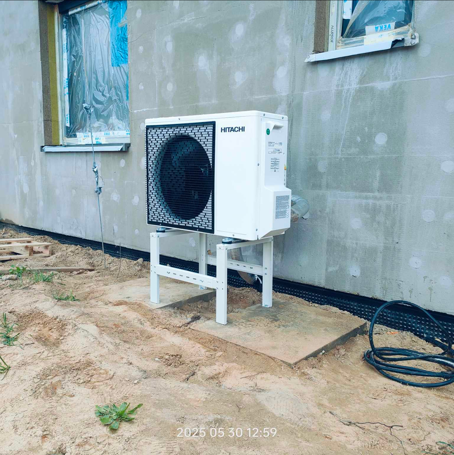 Biała pompa ciepła Hitachi na metalowym stelażu, zainstalowana przy ścianie budynku w trakcie budowy, z widocznym oknem zabezpieczonym folią.