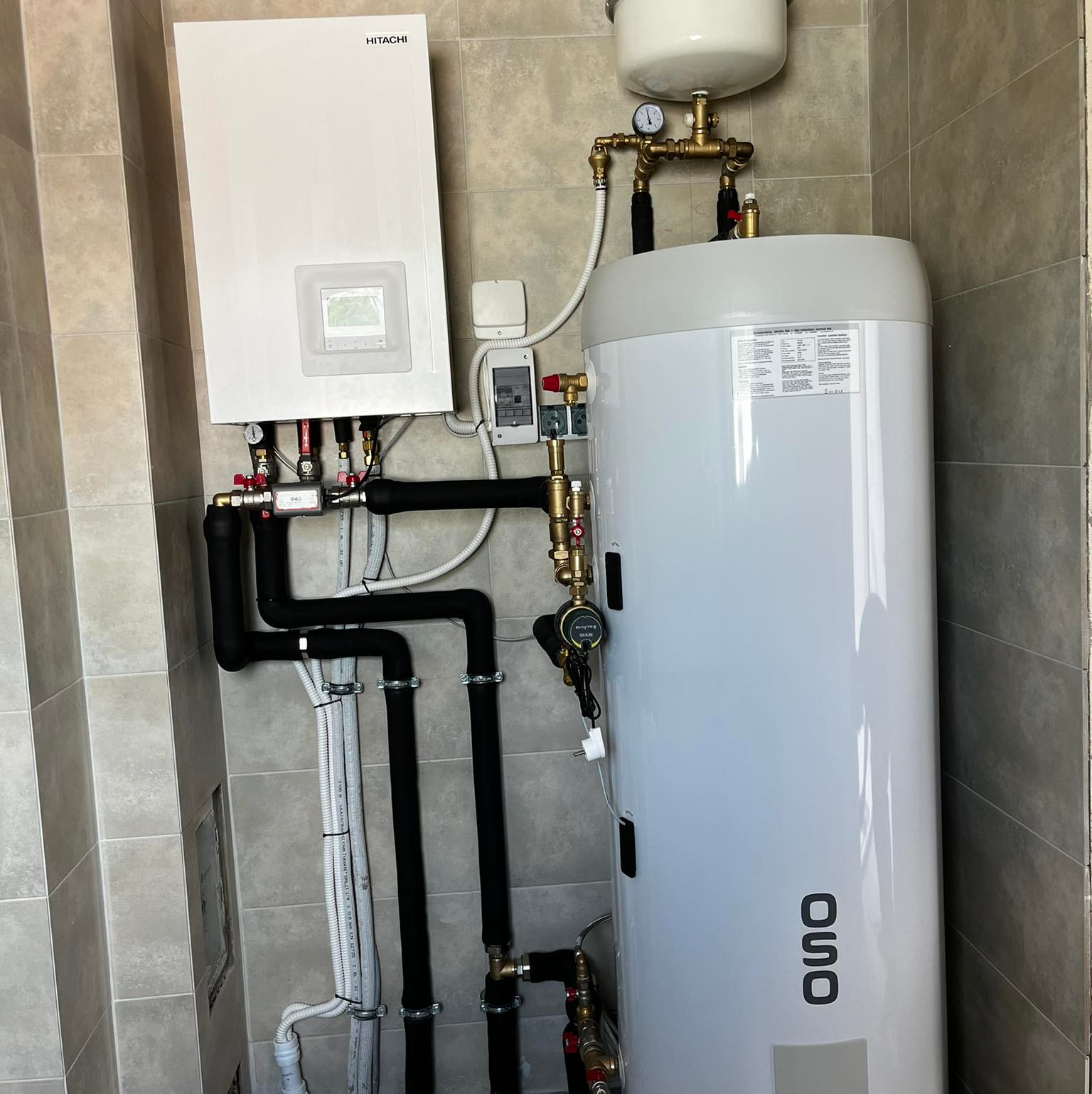 Biały bojler OSO i piec Hitachi na tle szarych płytek. Czarna izolacja rur. Instalacja grzewcza w kotłowni, po montażu. Widok z dołu.