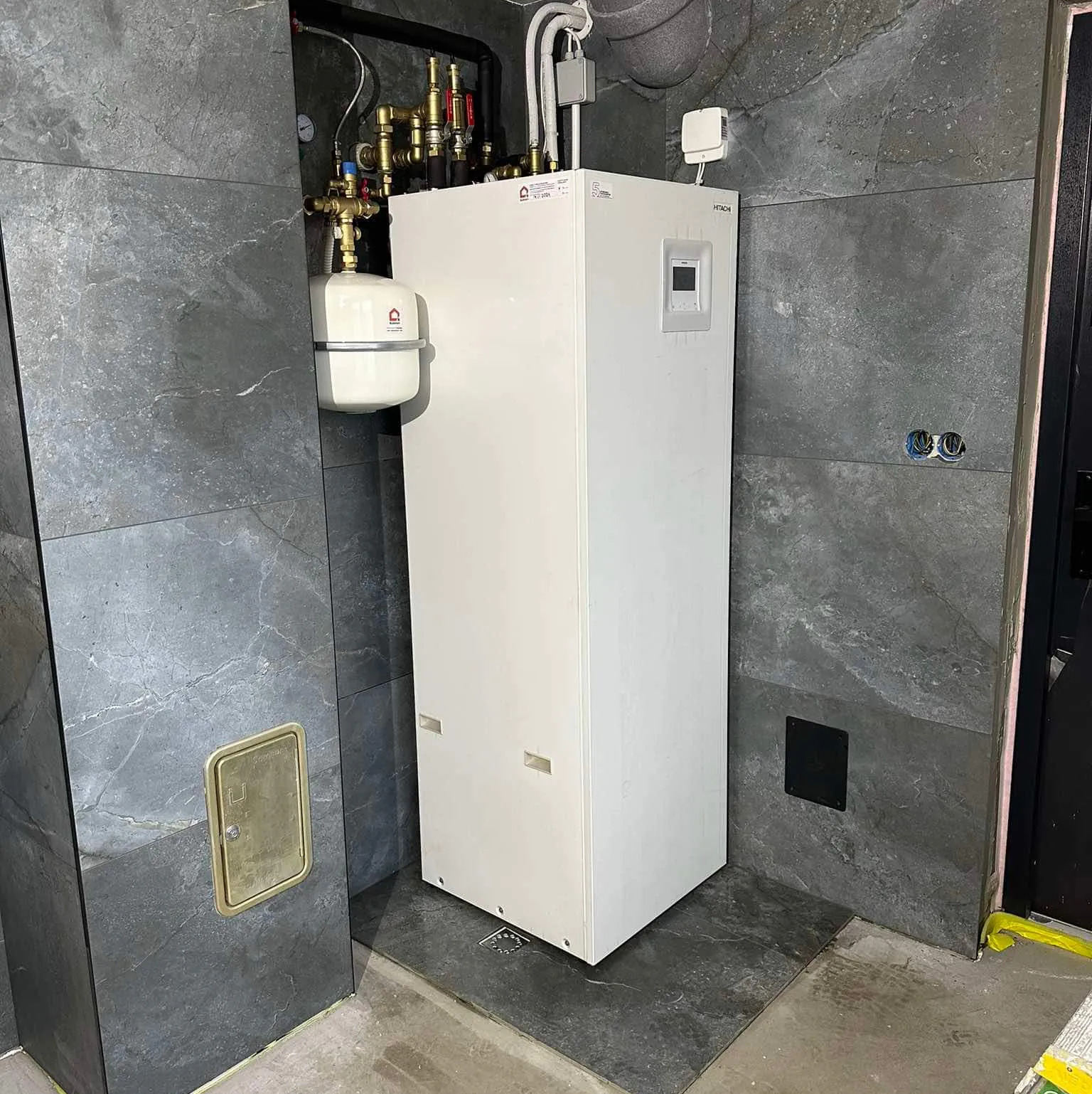 Biała pompa ciepła Hitachi, z osprzętem hydraulicznym, zainstalowana w pomieszczeniu wykończonym grafitową płytką imitującą kamień, widok z bliska.