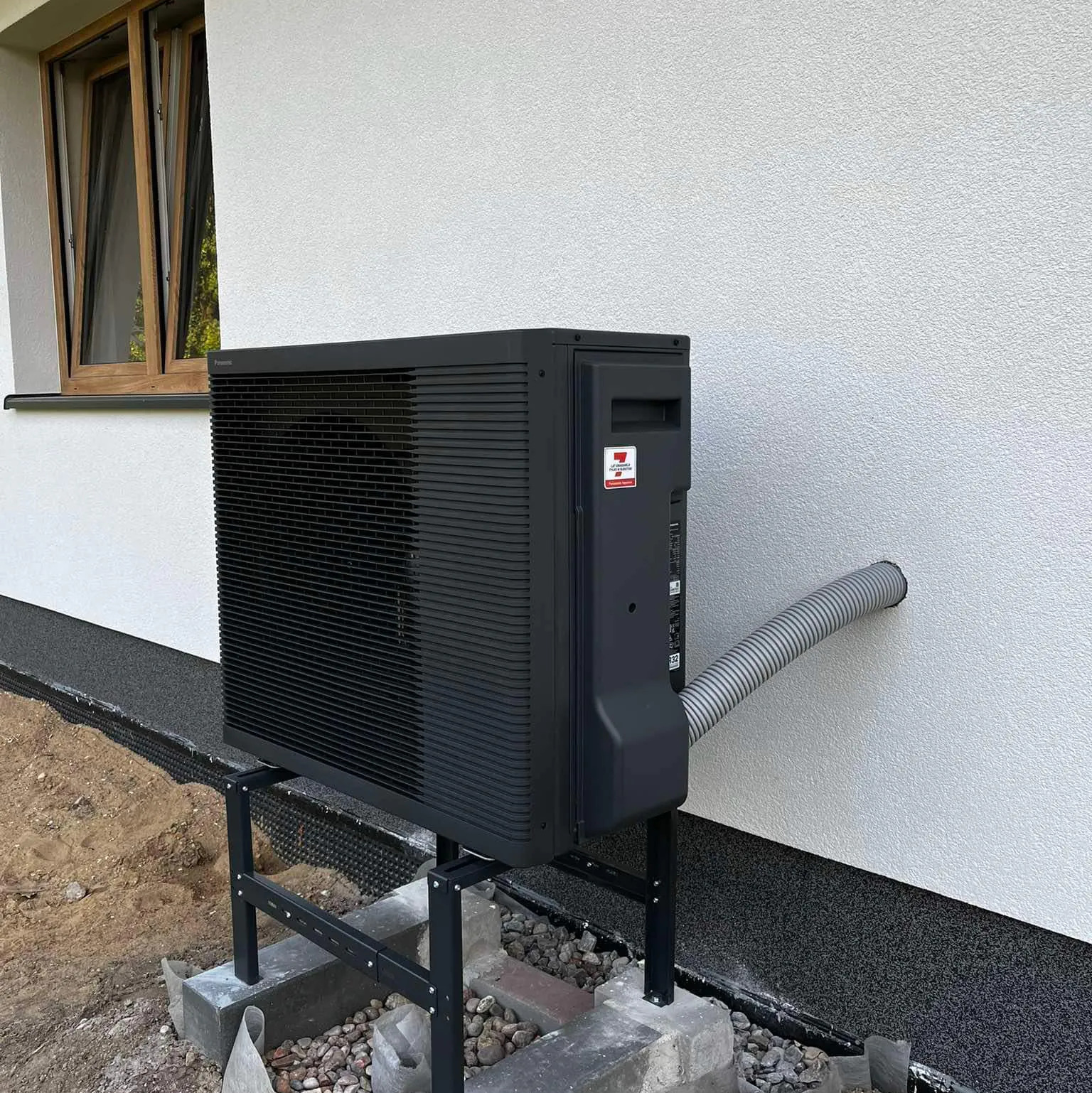 Czarna pompa ciepła zamontowana na zewnątrz budynku z widocznym przewodem wchodzącym w ścianę. Widoczne okno na górze i fragment fundamentu.